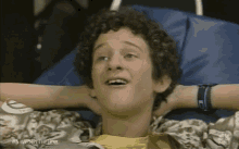 A young man with curly hair is laying in a bed with his hands behind his head 754281143634024